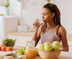 Woman drinking a glass of water, surrounded by healthy fruits and vegetables. 